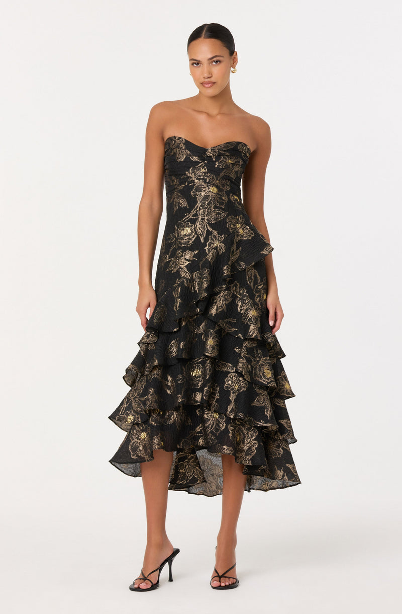 A woman is wearing a strapless, black dress with gold floral patterns. The dress features a fitted bodice and a layered, ruffled skirt that falls below the knees. She is also wearing black high-heeled sandals.