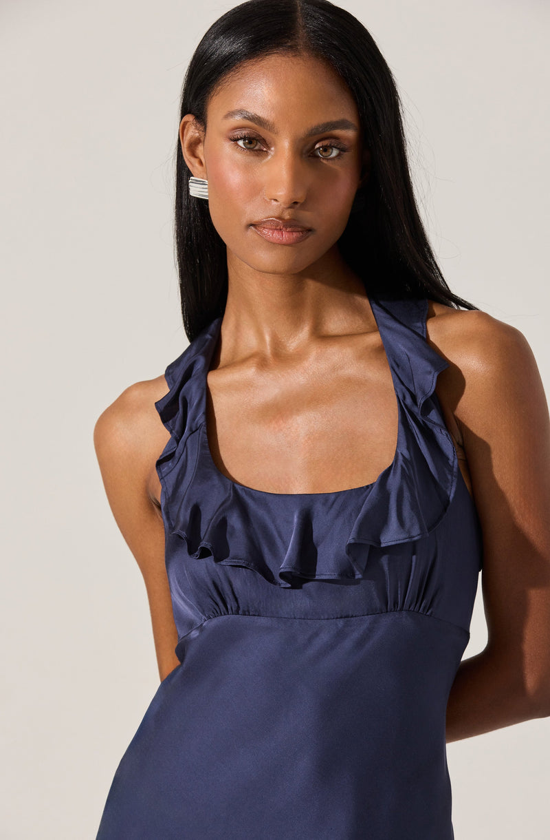 A woman is wearing a sleeveless, navy blue dress with ruffled details along the neckline and shoulder straps. The dress has a shiny satin-like fabric and a fitted bodice. She accessorizes with rectangular, silver earrings.