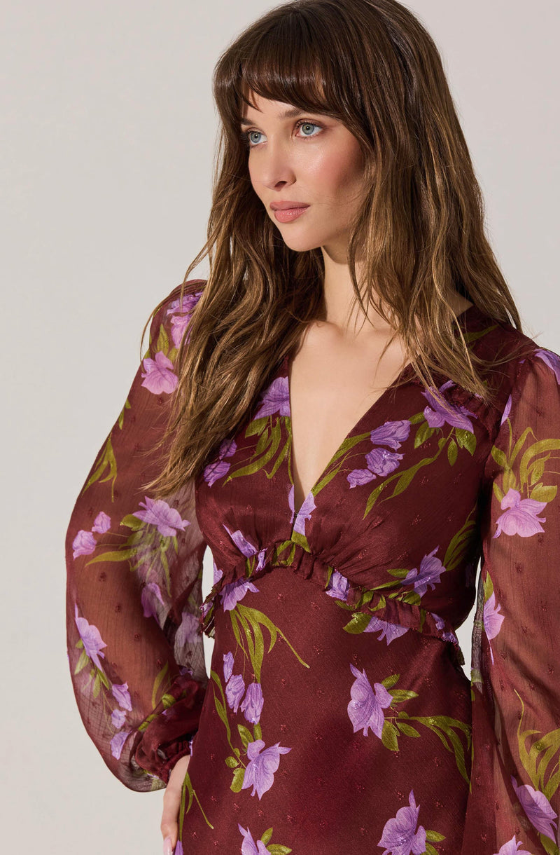 A woman is wearing a long-sleeved dress with a deep V neckline. The dress features a floral pattern with purple and pink flowers and green leaves on a burgundy background. The fabric appears lightweight and slightly sheer, and the dress has a gathered detail at the waist.