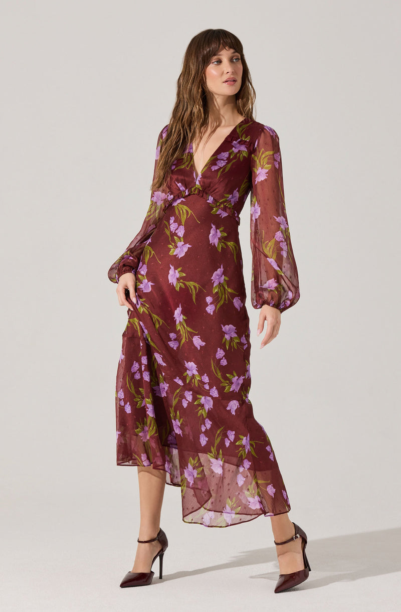 A woman is wearing a long, flowy dress with a deep V-neckline. The dress is sheer and has a maroon base color with a floral pattern of purple flowers and green leaves. The dress features long, slightly puffed sleeves gathered at the wrists and has a high slit on one side.