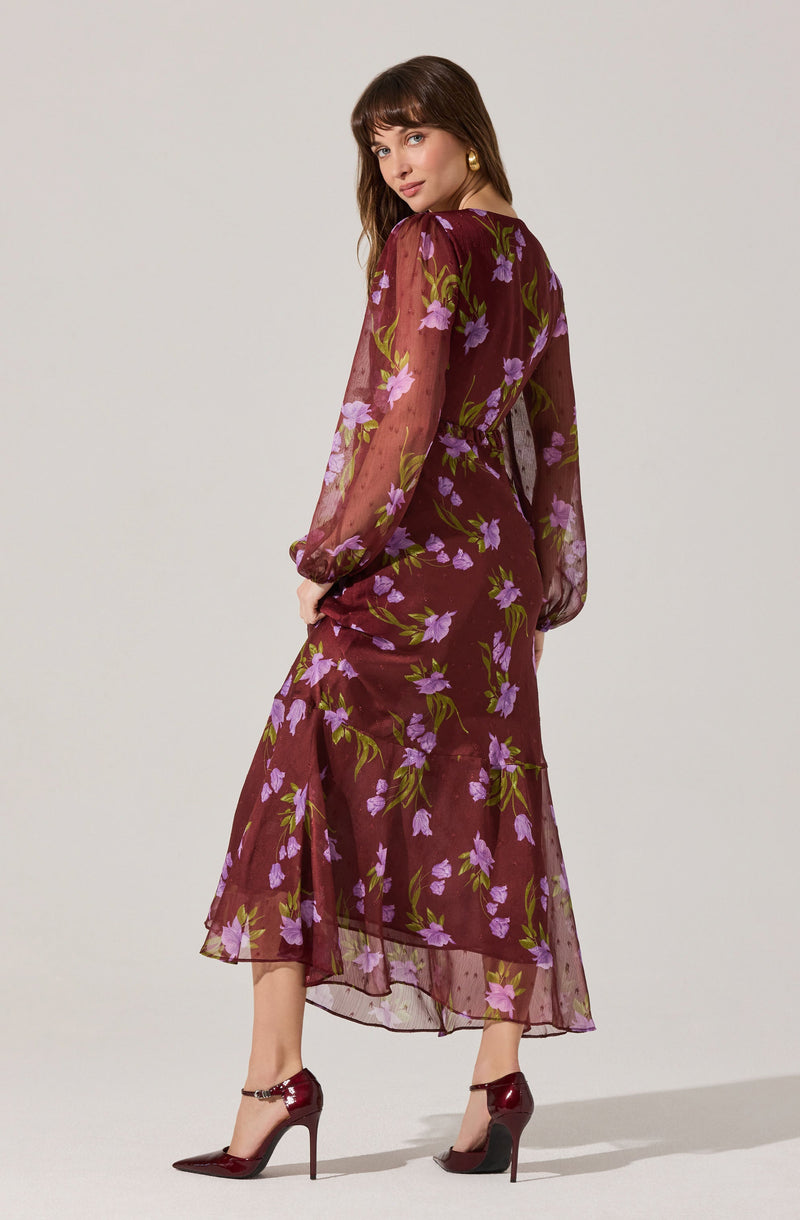 A woman is wearing a long, sheer burgundy dress with a floral print of purple flowers and green leaves. The dress has long, puffy sheer sleeves, a fitted waist, and a flowy skirt that falls to her ankles. She is also wearing burgundy high-heeled shoes with ankle straps.