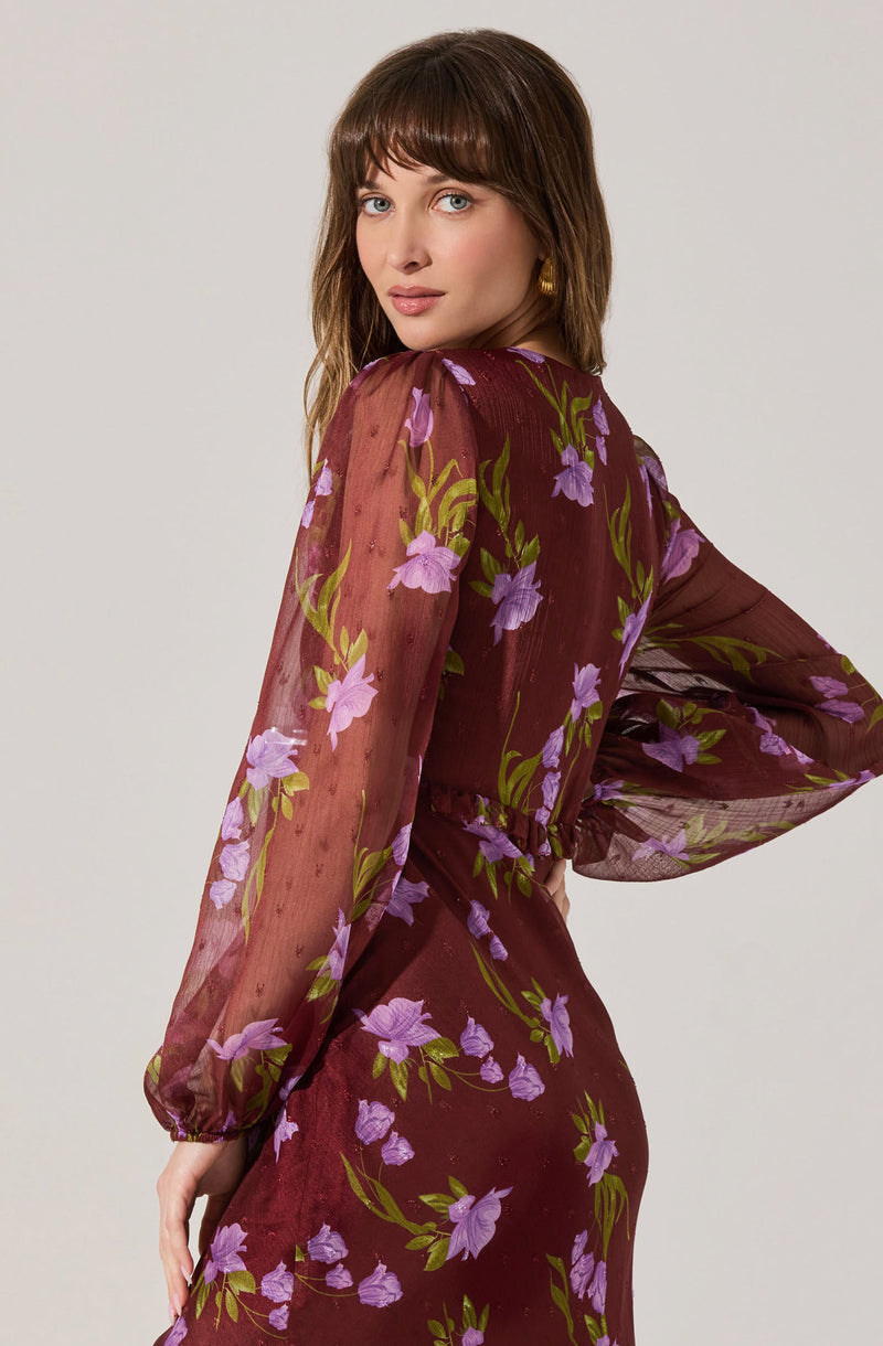 A woman is wearing a sheer, long-sleeved dress with a maroon background and a floral pattern of purple flowers with green leaves.