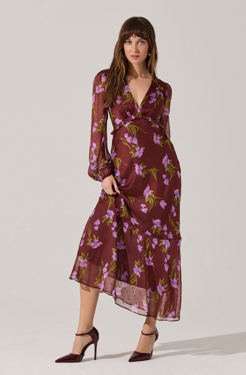 A woman is wearing a long, burgundy dress with a floral pattern of purple flowers and green leaves. The dress has long, semi-transparent sleeves with elastic cuffs, a V-neckline, and a gathered waist. The fabric is lightweight and flowy, reaching down to her ankles. She is also wearing burgundy high-heeled shoes.