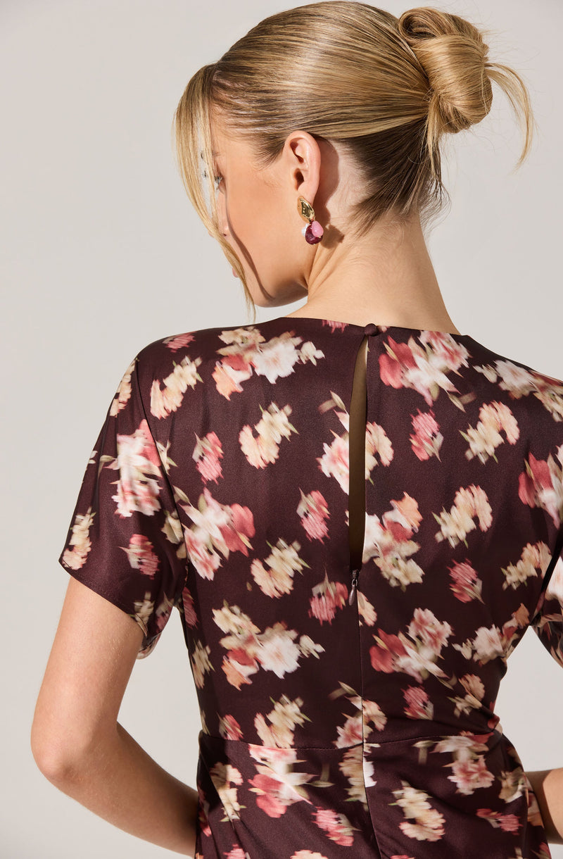 Back view of a woman wearing a dark-colored dress with a floral pattern featuring pink, white, and beige flowers. The dress has short sleeves and a vertical zipper running down the back. The fabric appears lightweight and slightly sheer. The woman has blonde hair styled in a low, loose bun and is wearing large, dangling earrings.