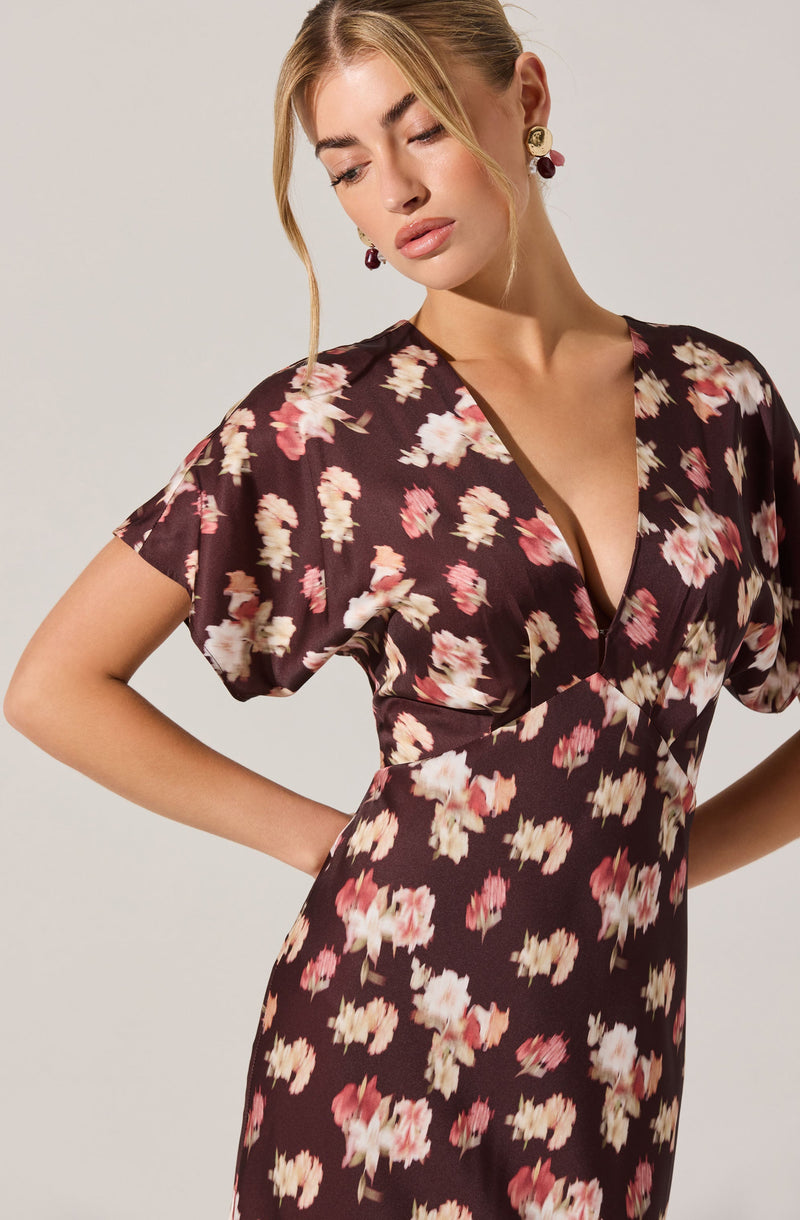 A woman is wearing a dark brown dress with a floral pattern featuring pink, cream, and white flowers. The dress has short, slightly puffy sleeves and a deep V neckline.