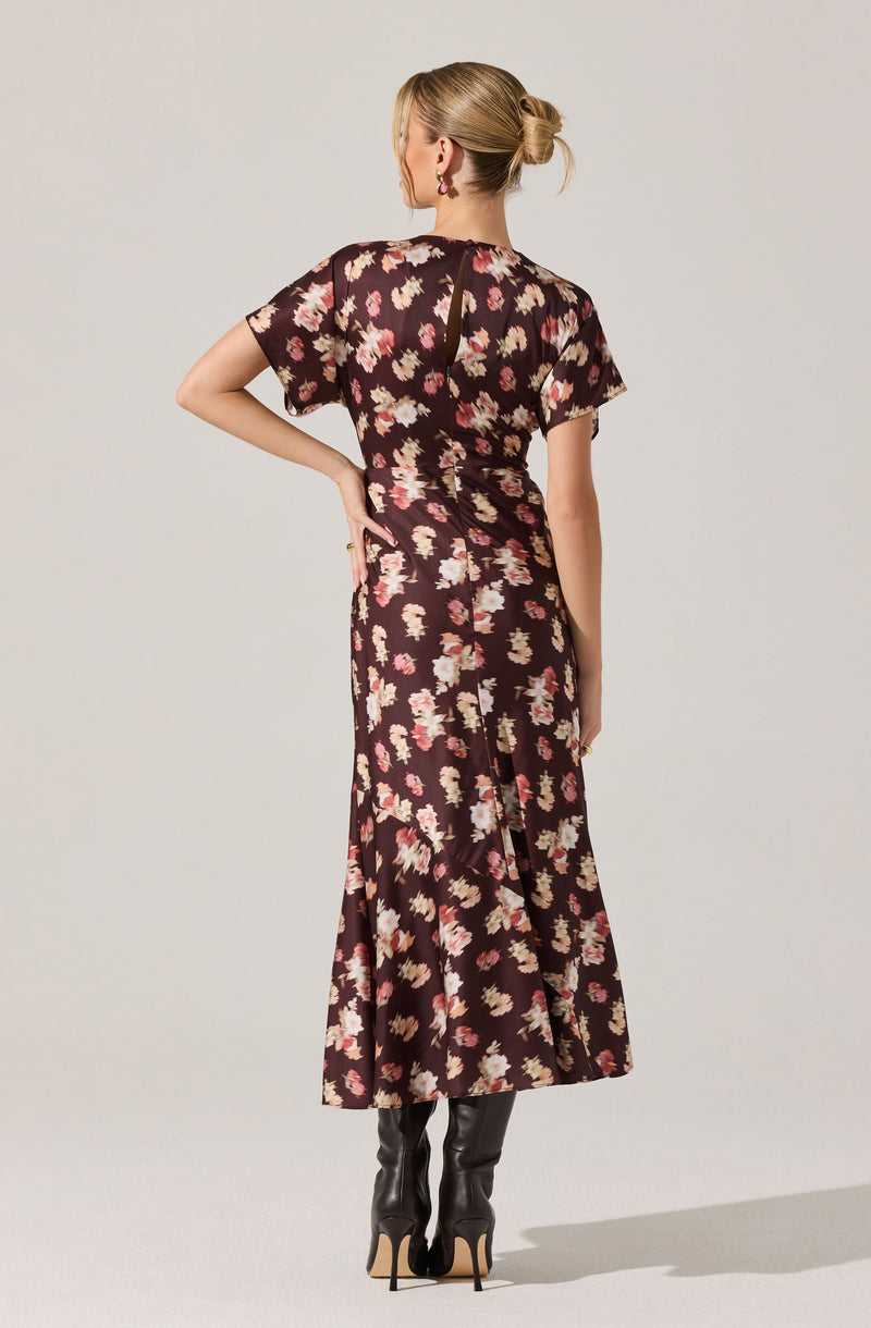 A woman is wearing a dark floral dress with pink and beige flower patterns. The dress has short sleeves and a round neckline, with a slit at the back. She is also wearing black knee-high boots with high heels.