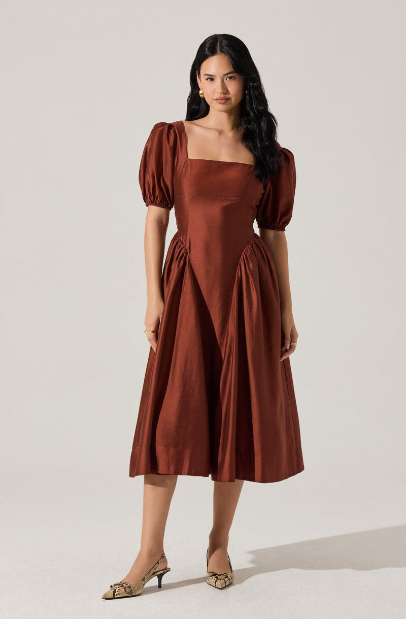 A woman wearing a mid-length, rust-colored dress with puffed short sleeves, a square neckline, and pleated details on the skirt. She is also wearing beige heels with a animal print pattern.