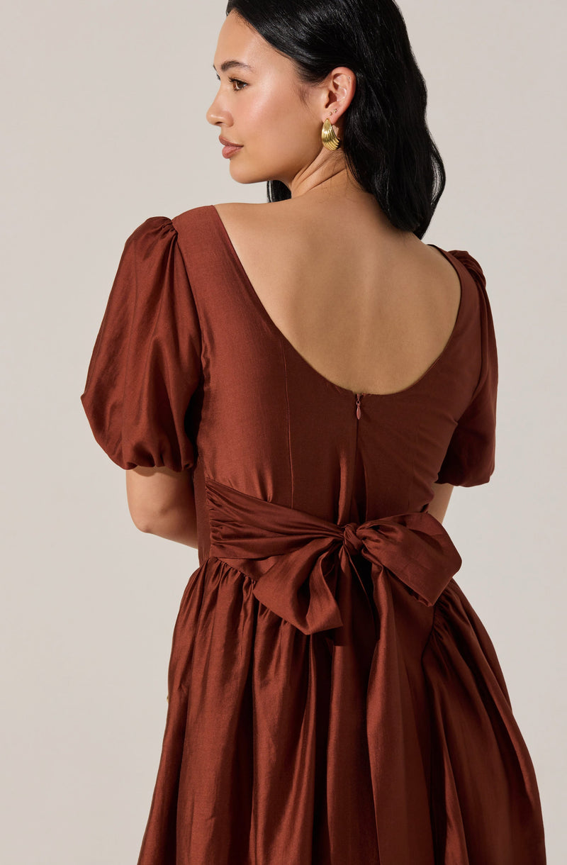A woman is wearing a bronze-colored dress made of a shiny, satin-like fabric. The dress features puffed short sleeves, a wide scoop neckline at the back, and a large, tied bow at the waist. The skirt is gathered and falls in soft pleats from the waist. She has dark, shoulder-length hair and is accessorized with large, gold hoop earrings.