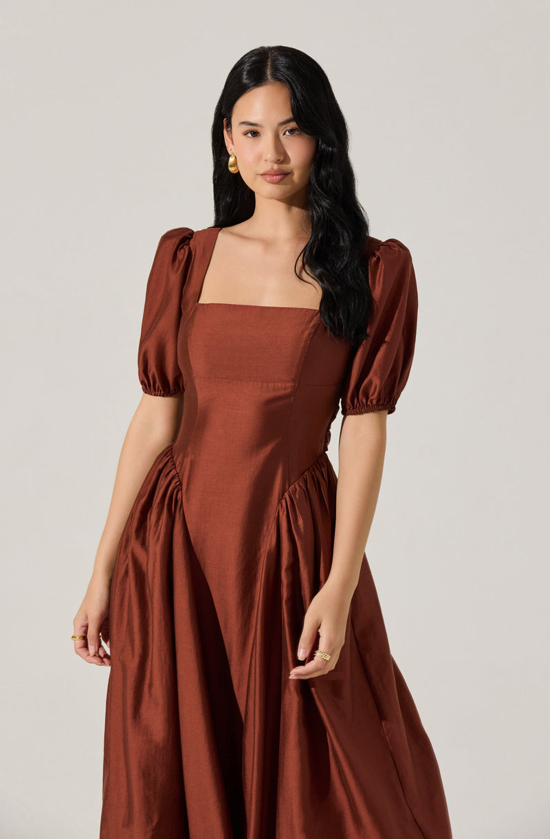 A woman is wearing a rust-colored gown with a fitted bodice and a square neckline. The dress has puffed short sleeves gathered at the ends, and the skirt is voluminous with gathered fabric at the sides. The material appears to be shiny and smooth, giving the dress an elegant look.