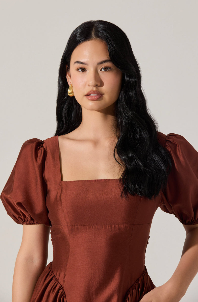 A woman wearing a satin dress in a rust color with puffed short sleeves, a square neckline, and a fitted bodice. She has long, wavy black hair and is accessorized with gold hoop earrings.