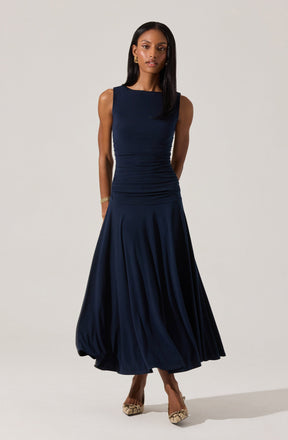 A woman is wearing a sleeveless, dark blue, midi-length dress with a fitted bodice that features ruching details. The skirt is full and gathered, creating a flowing silhouette. She accessorizes with gold earrings and a gold bracelet, and is wearing high-heeled shoes with a snakeskin pattern.
