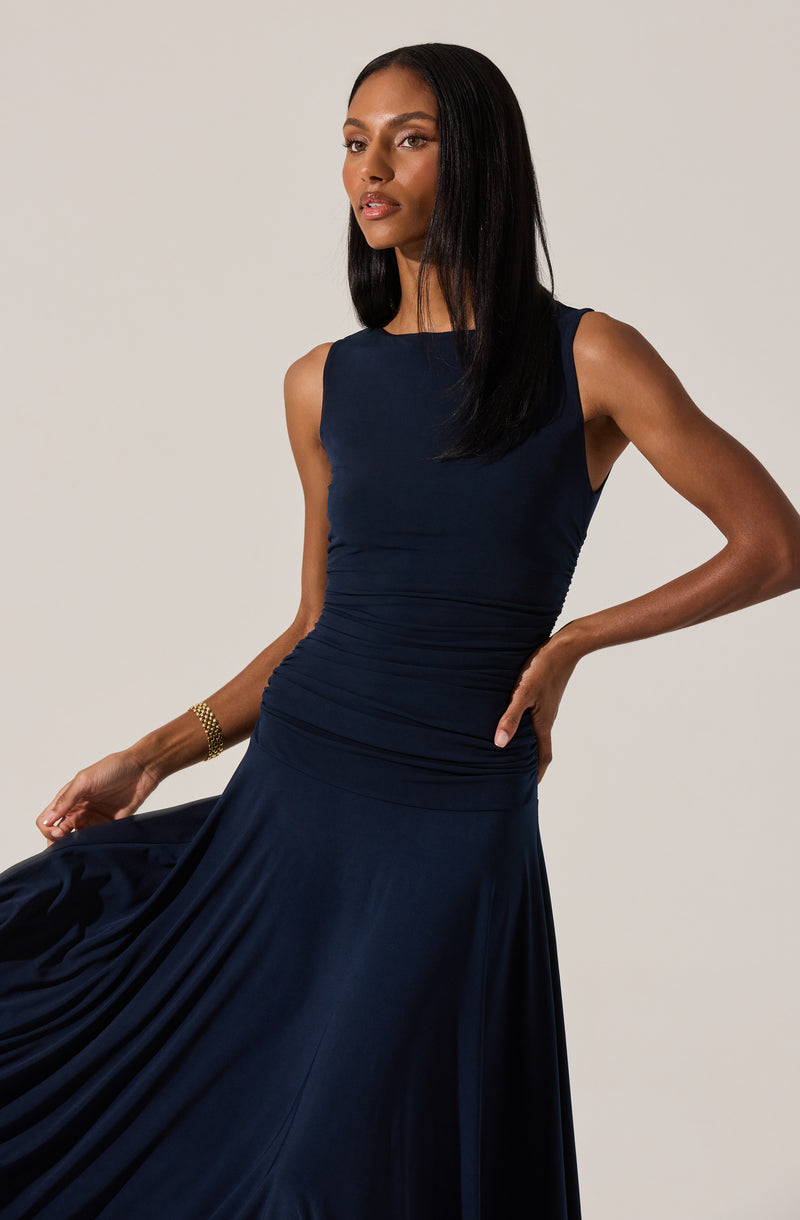 A woman is wearing a sleeveless, fitted navy blue dress with a high neckline. The dress is ruched around the waist and hips, with a flowing skirt made of a smooth, shiny fabric. She has a gold bracelet on her wrist.