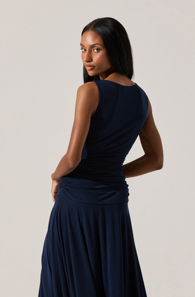 A woman is wearing a sleeveless, dark navy blue dress with a fitted bodice and a draped, flowing skirt. The dress has a round neckline and appears to be made of a smooth, slightly shiny fabric. She is standing with her back partially turned, giving a side and rear view of the dress.