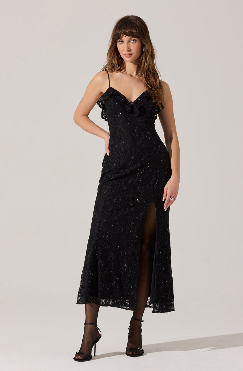 A woman is wearing a long, black, glittery evening gown with thin spaghetti straps and a ruffled neckline. The dress features a high slit in the front, revealing her legs and black sheer stockings. She is also wearing black high-heeled sandals with ankle straps.