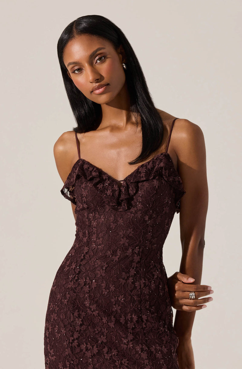 A woman is wearing a fitted, sleeveless dress made of burgundy lace fabric. The dress features delicate spaghetti straps and a ruffled lace detail along the neckline. The lace pattern is intricate and floral, adding texture and elegance to the garment.