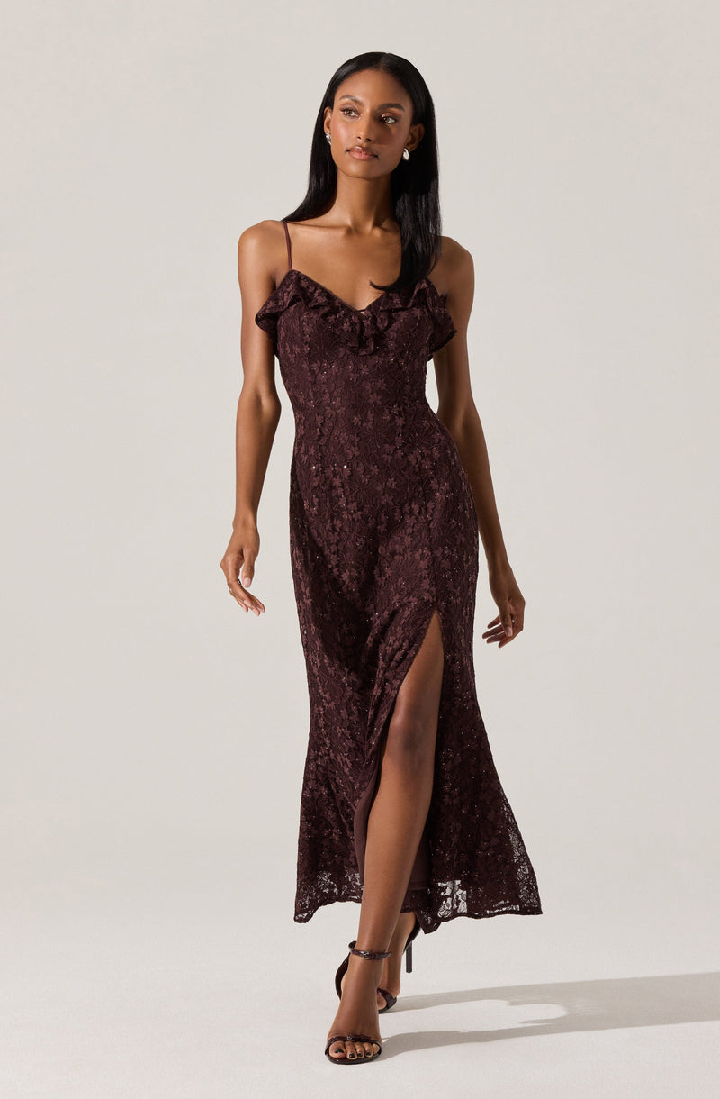 A woman is wearing a long, burgundy lace dress with thin spaghetti straps. The dress features ruffle details at the neckline and has a high slit up one side, revealing her leg. She is wearing strappy high-heeled sandals.