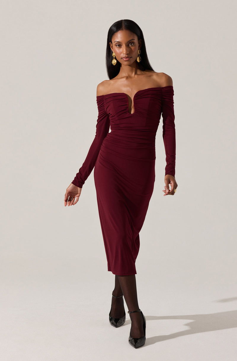 A woman wearing an elegant, off-the-shoulder, long-sleeve burgundy dress with ruched detailing across the bodice and sleeves. The dress has a fitted silhouette that falls to mid-calf length. She pairs the outfit with black tights, black heels, and gold earrings.