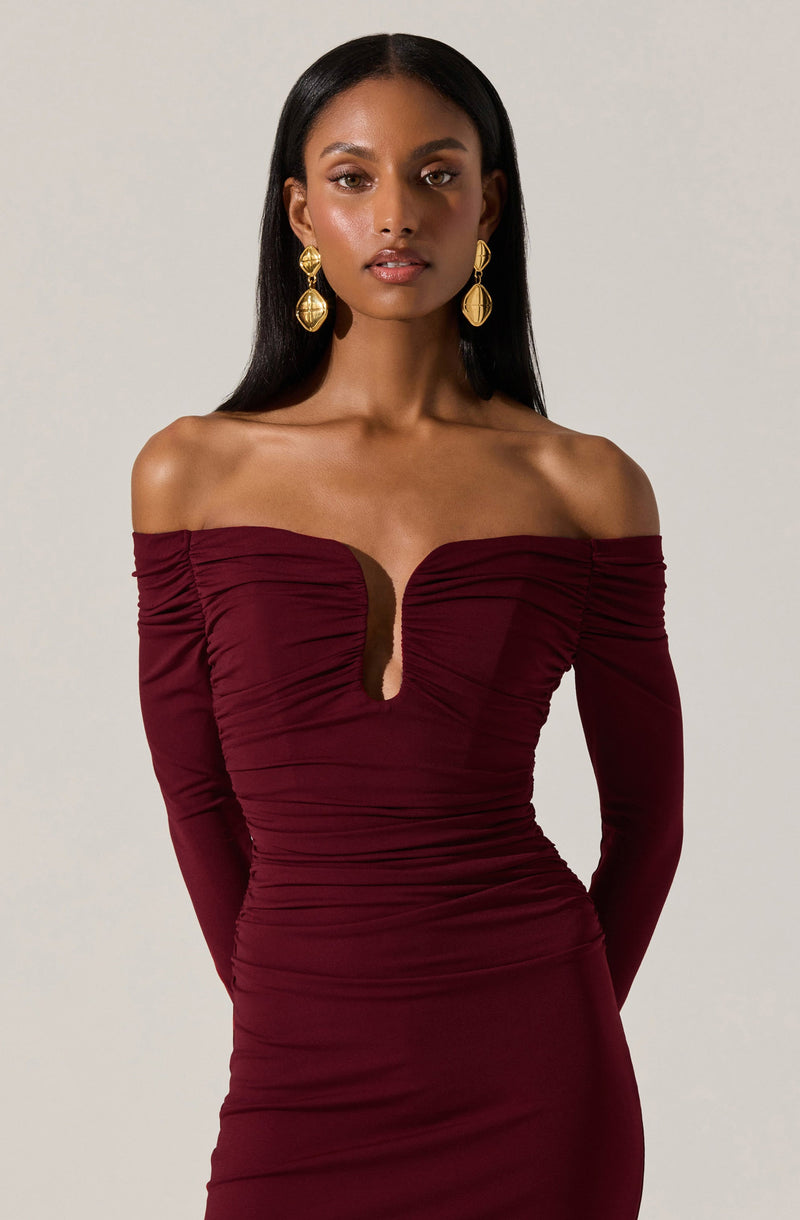 A woman is wearing an off-the-shoulder burgundy dress with long sleeves. The dress features gathered fabric throughout and a keyhole cutout at the chest. She is accessorized with large, gold, dangle earrings.