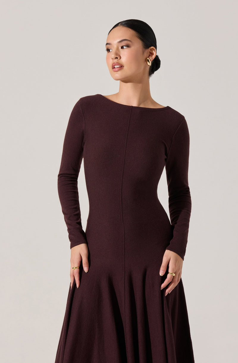 A woman is wearing a fitted, long-sleeved, dark burgundy dress with a round neckline. The dress features a subtle vertical seam down the front and a pleated, flowing skirt that extends to the floor. She has her hair tied back and is accessorized with gold hoop earrings and gold rings.