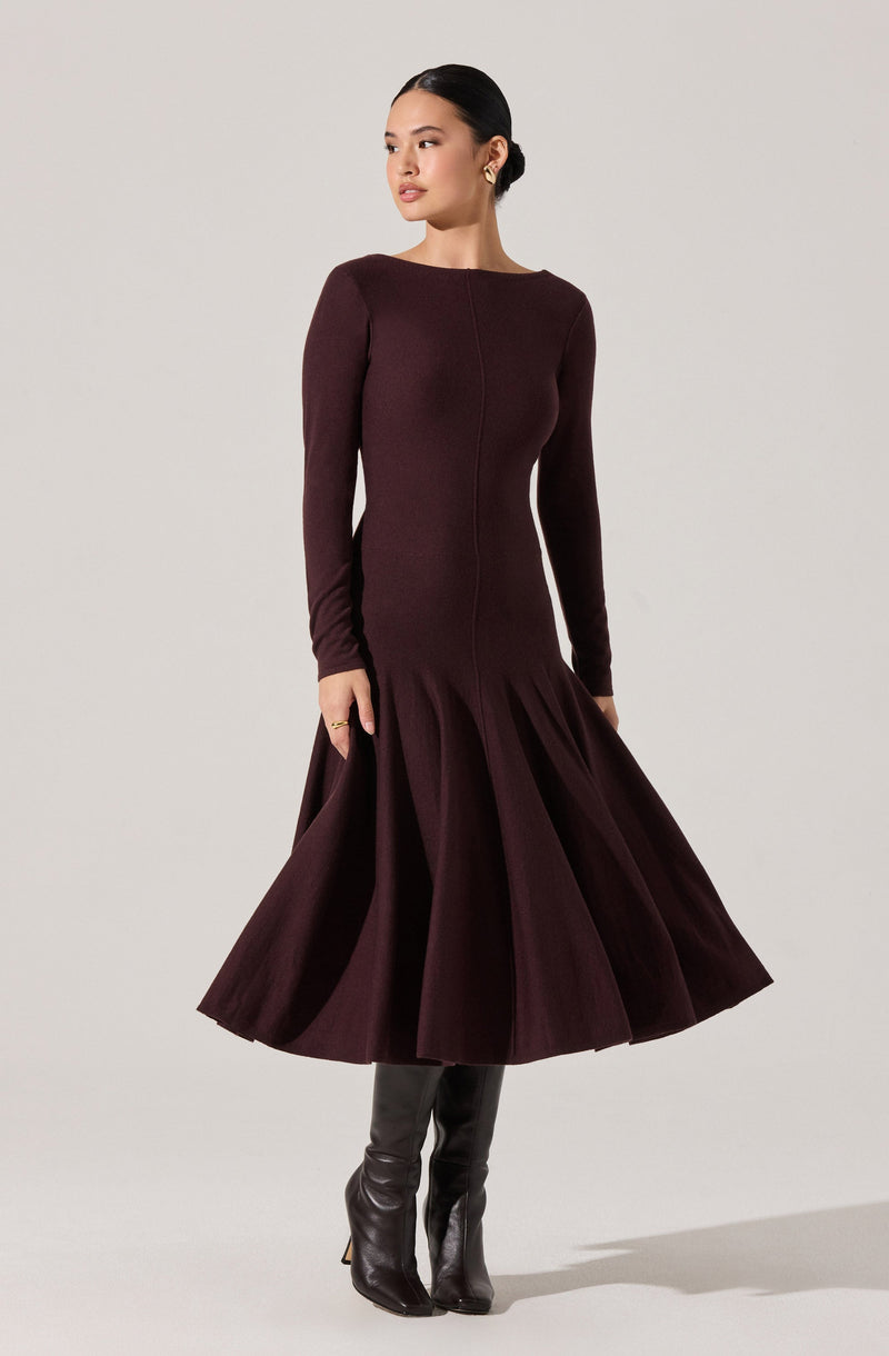 A woman is wearing a long-sleeved, fitted burgundy dress with a boat neckline. The dress has a flared, pleated skirt that extends to mid-calf length. She pairs the outfit with tall, black leather boots.