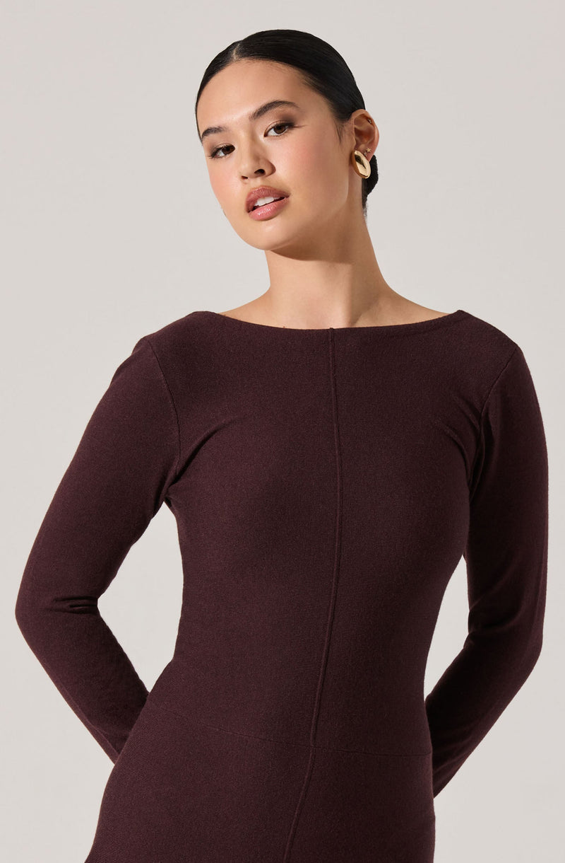A woman is wearing a long-sleeved, form-fitting dress in a dark burgundy color. The dress has a simple, elegant design with a round neckline and visible seam details running vertically down the front. She has dark hair pulled back and is accessorizing with large, rounded gold earrings.