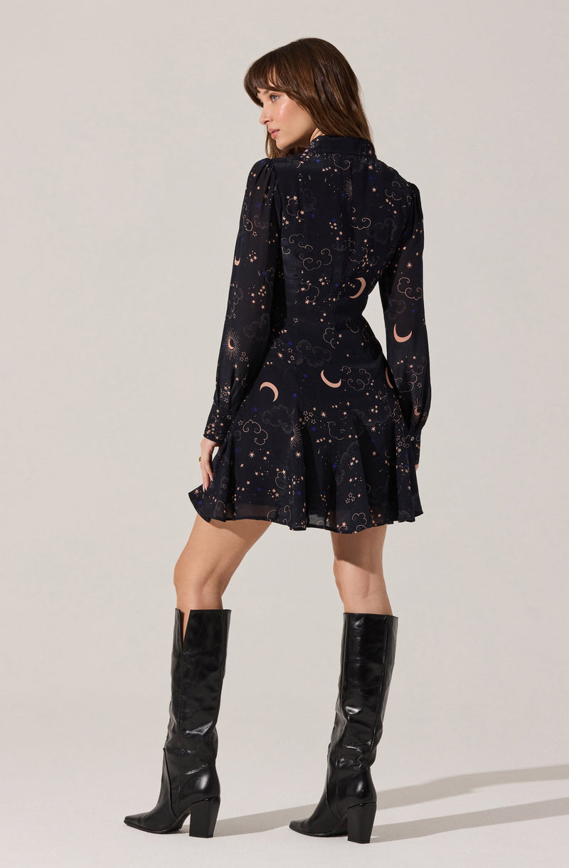A woman is wearing a dark dress with a celestial pattern featuring moons and stars. The dress has long sheer sleeves, a fitted waist, and a flared skirt. She pairs the dress with tall, shiny black knee-high boots with block heels.