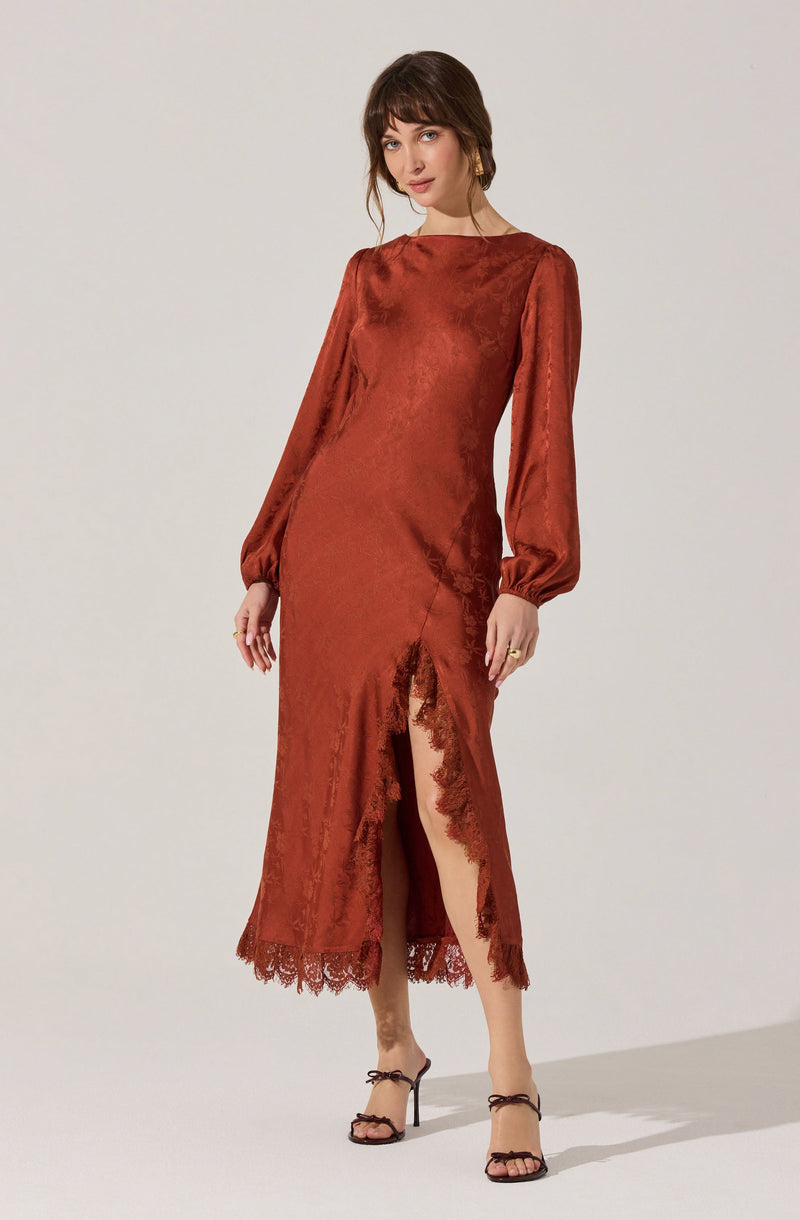 A woman is wearing a long, rust-colored satin dress with a subtle sheen. The dress features long, puffed sleeves gathered at the wrists, a fitted bodice, and a high neckline. It has a front slit that extends from the hem to mid-thigh, adorned with delicate lace trim along the edges of the slit and hemline. The dress has a smooth texture with a slight crinkled effect and appears to be designed for an elegant occasion. She completes her outfit with open-toe high-heeled sandals with ankle straps tied in bows.