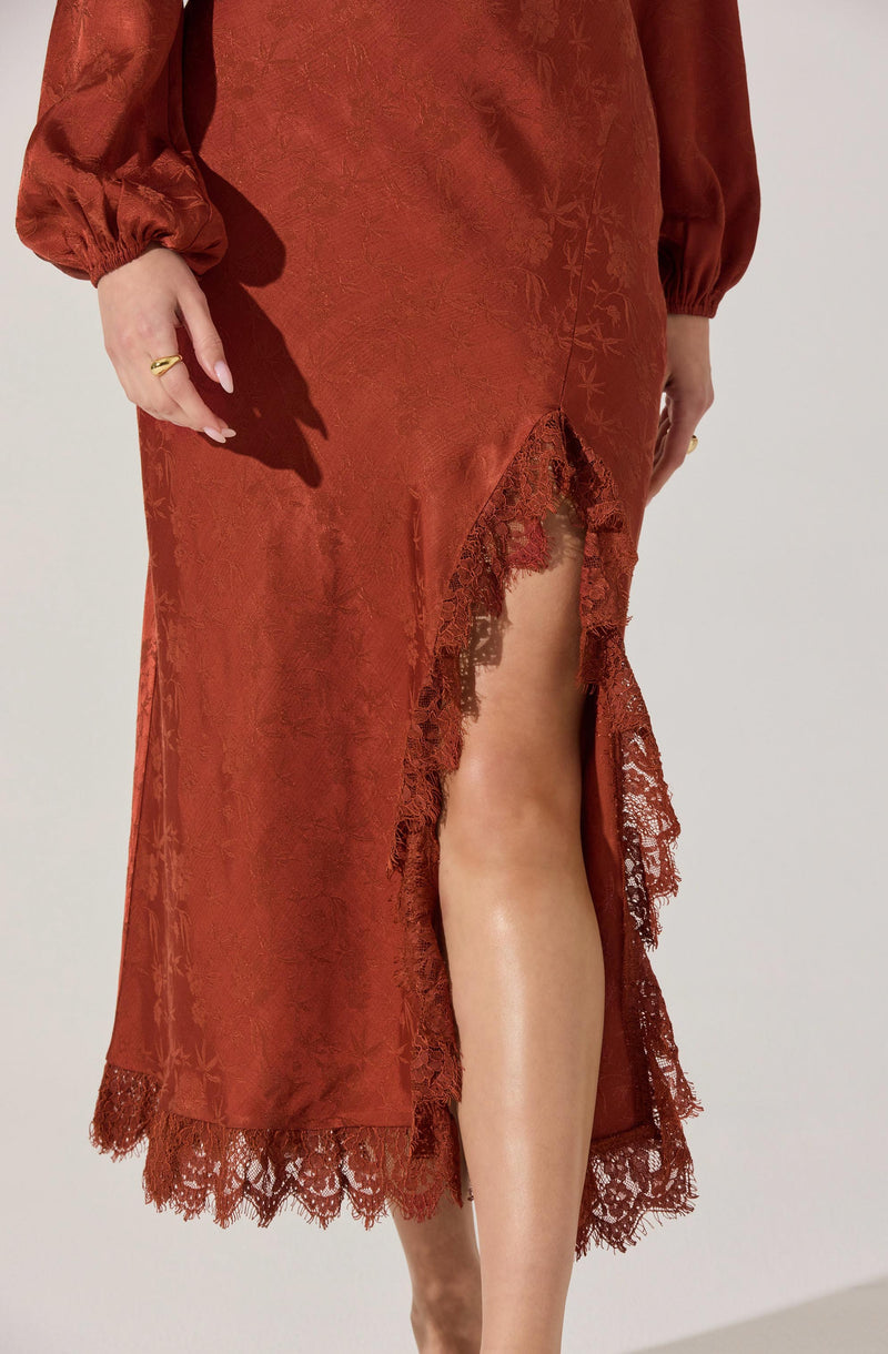 A woman is wearing a long, rust-colored satin dress with floral patterns. The dress has long, puffed sleeves gathered at the cuffs and features a high slit on the front, revealing her leg. The hemline and side slit are decorated with intricate lace trim that matches the color of the dress.