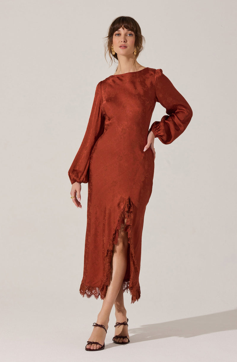 A woman is wearing a long, rust-colored satin dress with a subtle sheen. The dress has long, puffed sleeves gathered at the wrists and features a front slit with lace trim at the hem. She is also wearing black strappy high-heeled sandals.
