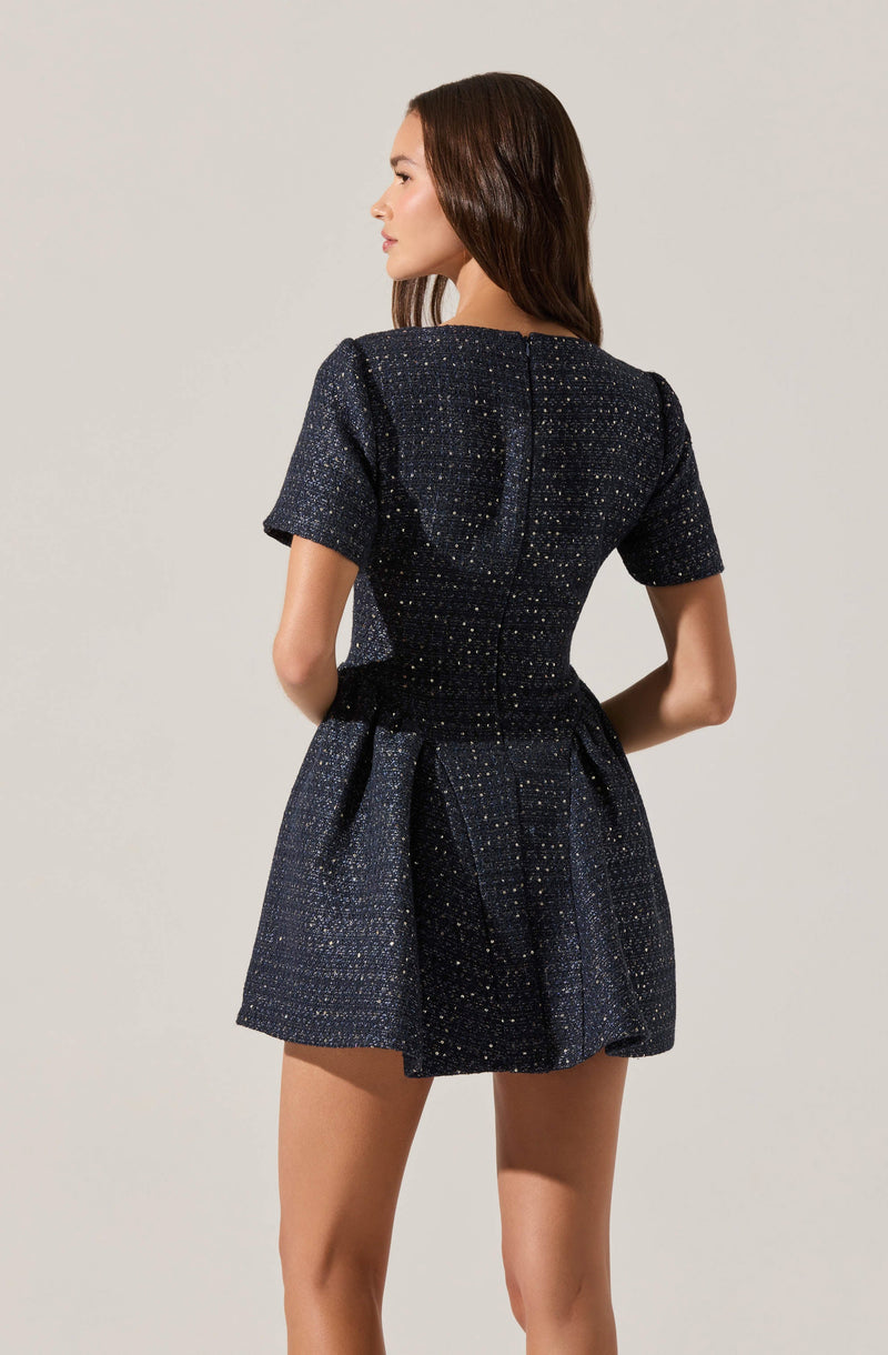 A woman is wearing a dark, textured, short-sleeved dress with a subtle shimmer or sparkle. The dress has a fitted bodice that cinches at the waist and a flared skirt that falls above the knees. The fabric appears to have a pattern of tiny, light-colored specks evenly distributed throughout.