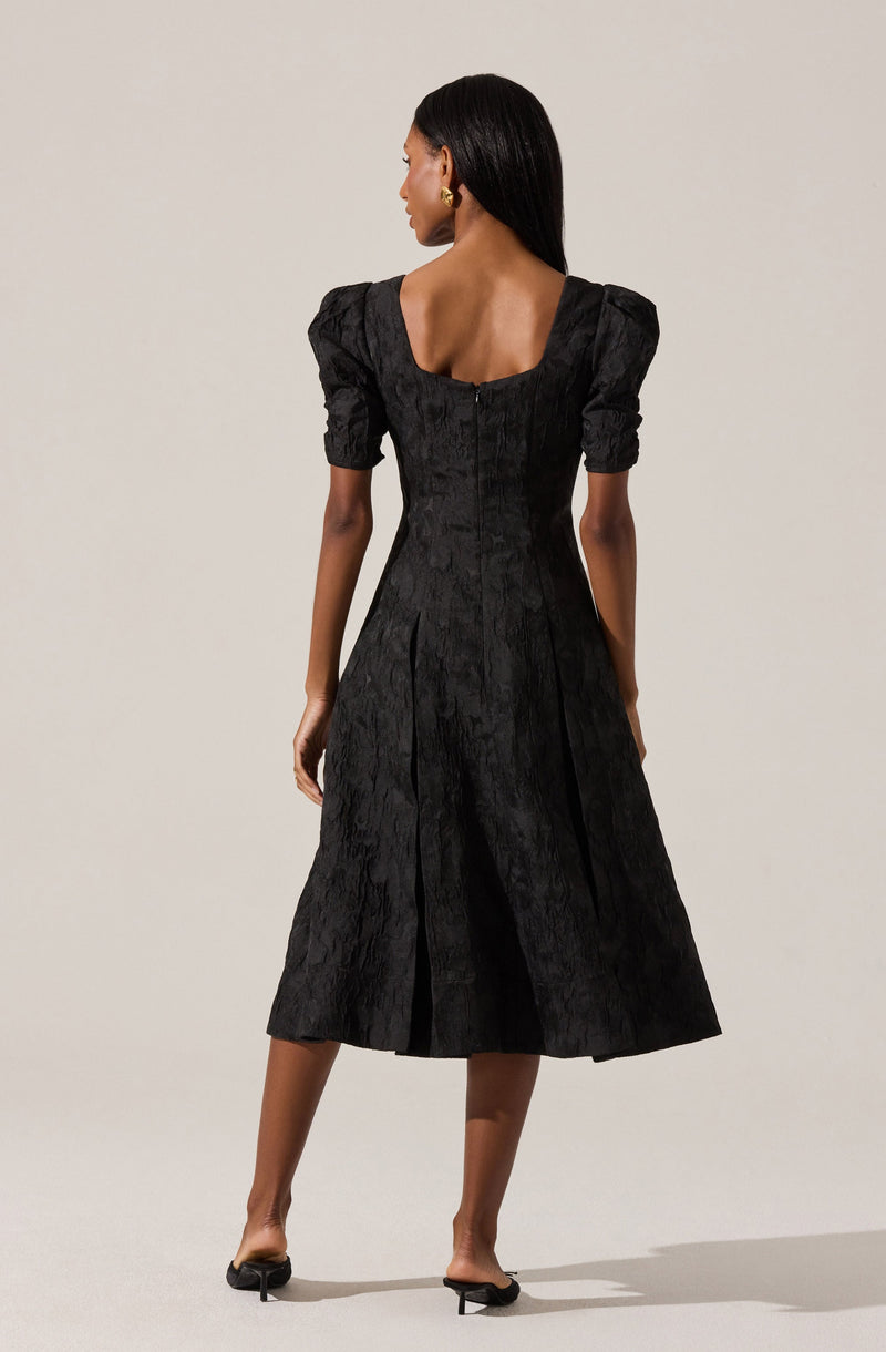 A woman is wearing a black dress with a textured, shiny fabric that has a subtle floral or damask pattern. The dress features a fitted bodice with a wide, square neckline, puffed short sleeves, and a full, midi-length skirt with pleats. She is also wearing small, gold hoop earrings and black, open-toed, heeled sandals.