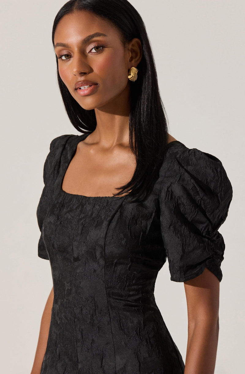 A woman is wearing a black dress made of textured, slightly shiny fabric with a subtle pattern. The dress has a square neckline and puffed short sleeves that gather at the shoulder and slightly below the elbow. She is accessorized with large gold earrings.