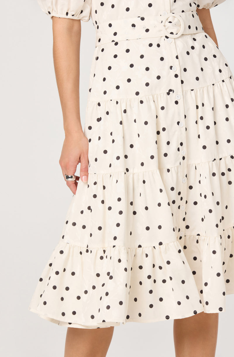 A woman is wearing a cream-colored, tiered midi dress with black polka dots. The dress features puffed short sleeves gathered at the cuffs and a matching fabric belt with a circular buckle at the waist. The skirt portion has horizontal seams creating ruffled tiers.
