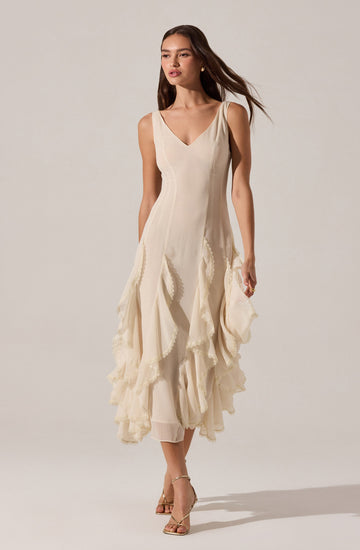 The dress is sleeveless with wide shoulder straps and a V-shaped neckline. It is made of a light, flowing fabric in an off-white or cream color. The dress features vertical seam lines on the bodice and has a layered, ruffled skirt with textured, scalloped edges, creating a tiered effect that extends to mid-calf length. The overall style is elegant and feminine.