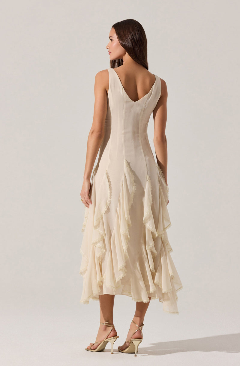 A woman is wearing an elegant sleeveless dress with a fitted bodice and a deep V neckline at the back. The dress is made of a light, cream-colored fabric with cascading, layered ruffles that run down from the waist to the hem, creating a textured, flowing appearance. The dress is midi-length with a slightly uneven hemline, and she is wearing strappy high-heeled sandals.