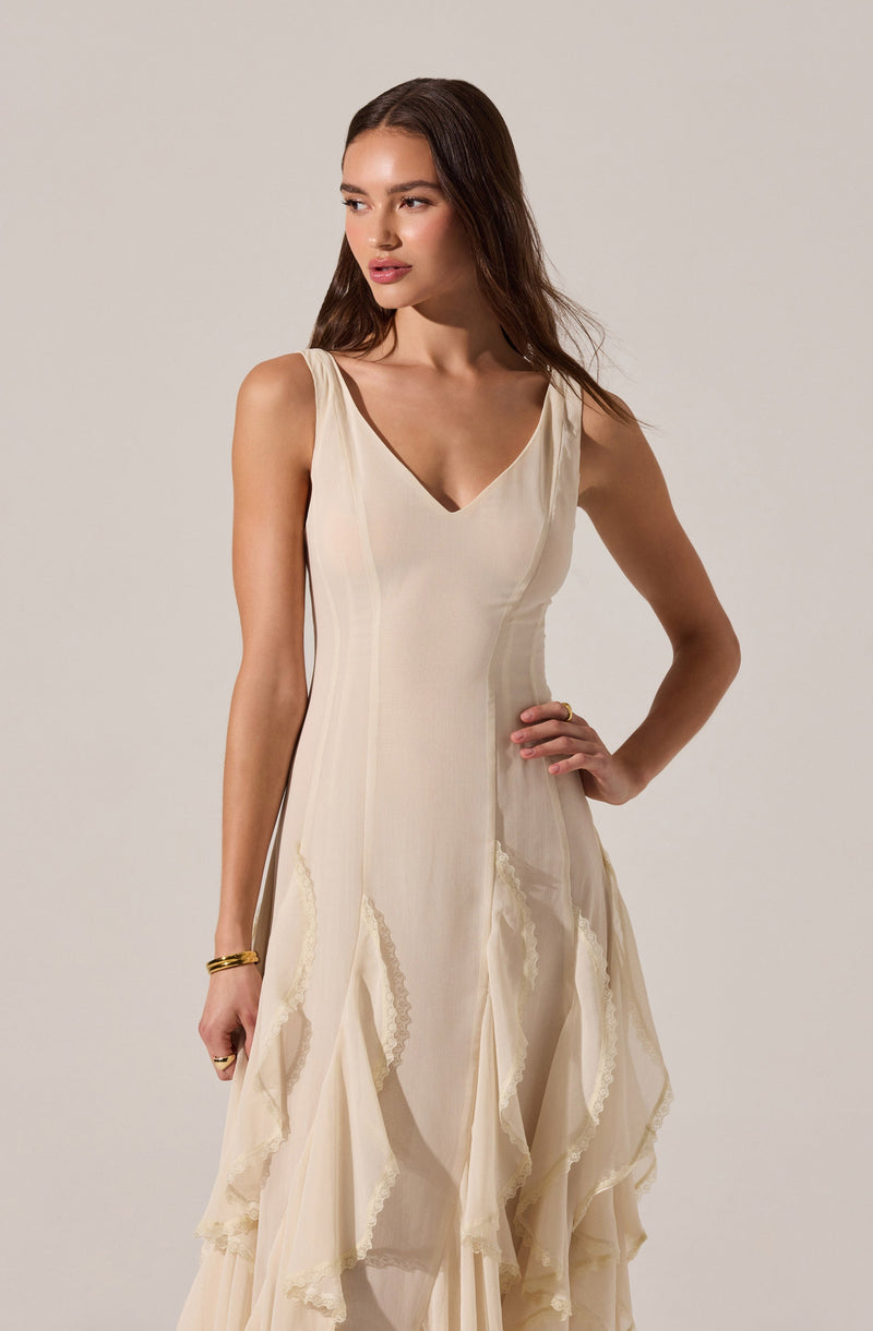 A woman is wearing an elegant sleeveless cream-colored dress with a V-neckline. The dress features vertical seam details on the bodice and layered, ruffled fabric accents with lace trim along the edges starting from the waist down to the hem, creating a flowing, textured look. She has minimal accessories, including a gold bracelet and ring, and her long brown hair is styled in loose waves.