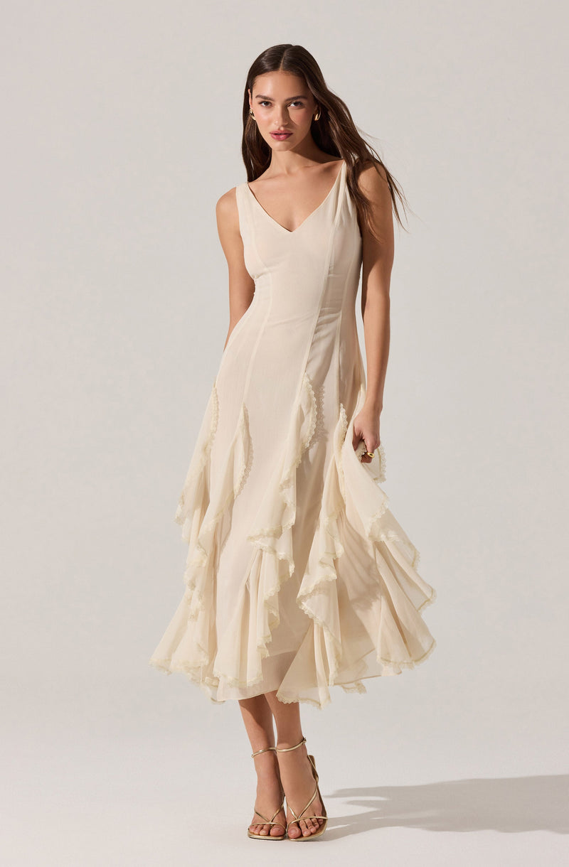 A woman is wearing a sleeveless, cream-colored dress with a V-neckline. The dress features layered, ruffled fabric panels that create a textured, flowing look. She has on strappy high-heeled sandals in a metallic or neutral tone.