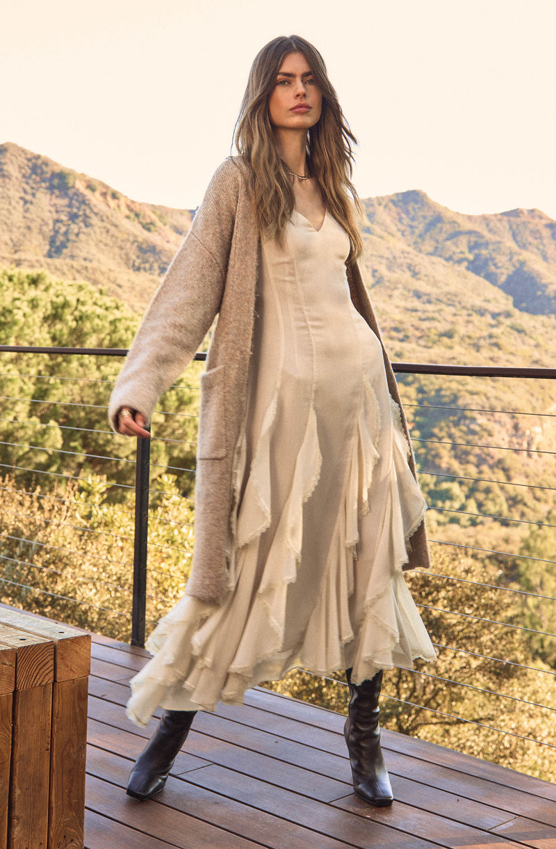 A woman is wearing a long, flowing cream-colored dress with layered, ruffled fabric details. She has on a beige, textured, open-front cardigan that extends to her knees. She completes her outfit with black knee-high boots.