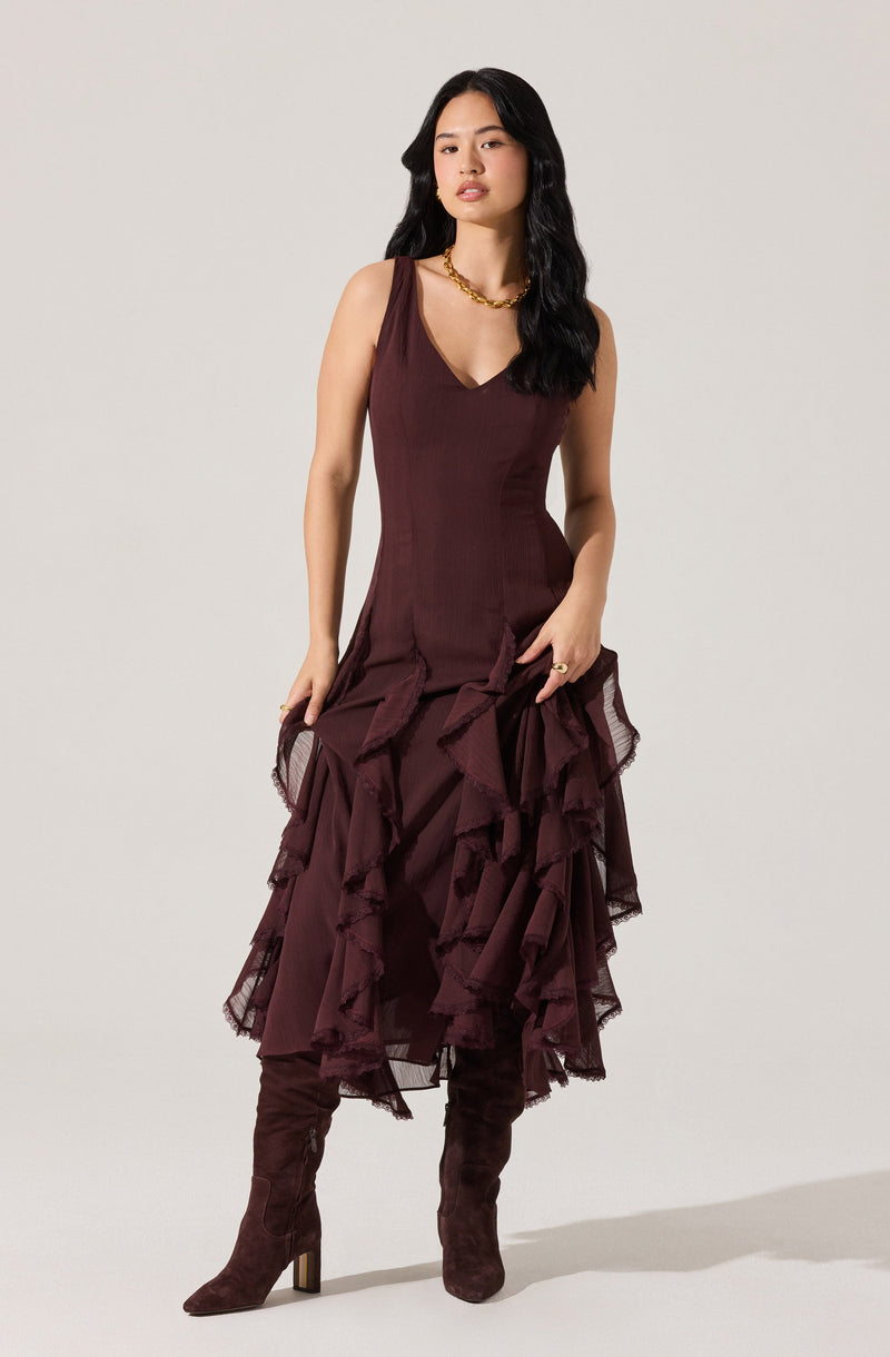 A woman is wearing a sleeveless, deep V-neck, dark burgundy dress with a fitted bodice. The skirt features multiple layers of ruffled, sheer fabric that cascade asymmetrically down to mid-calf length. She has accessorized with a chunky gold chain necklace and wears dark brown, high-heeled, knee-high boots.