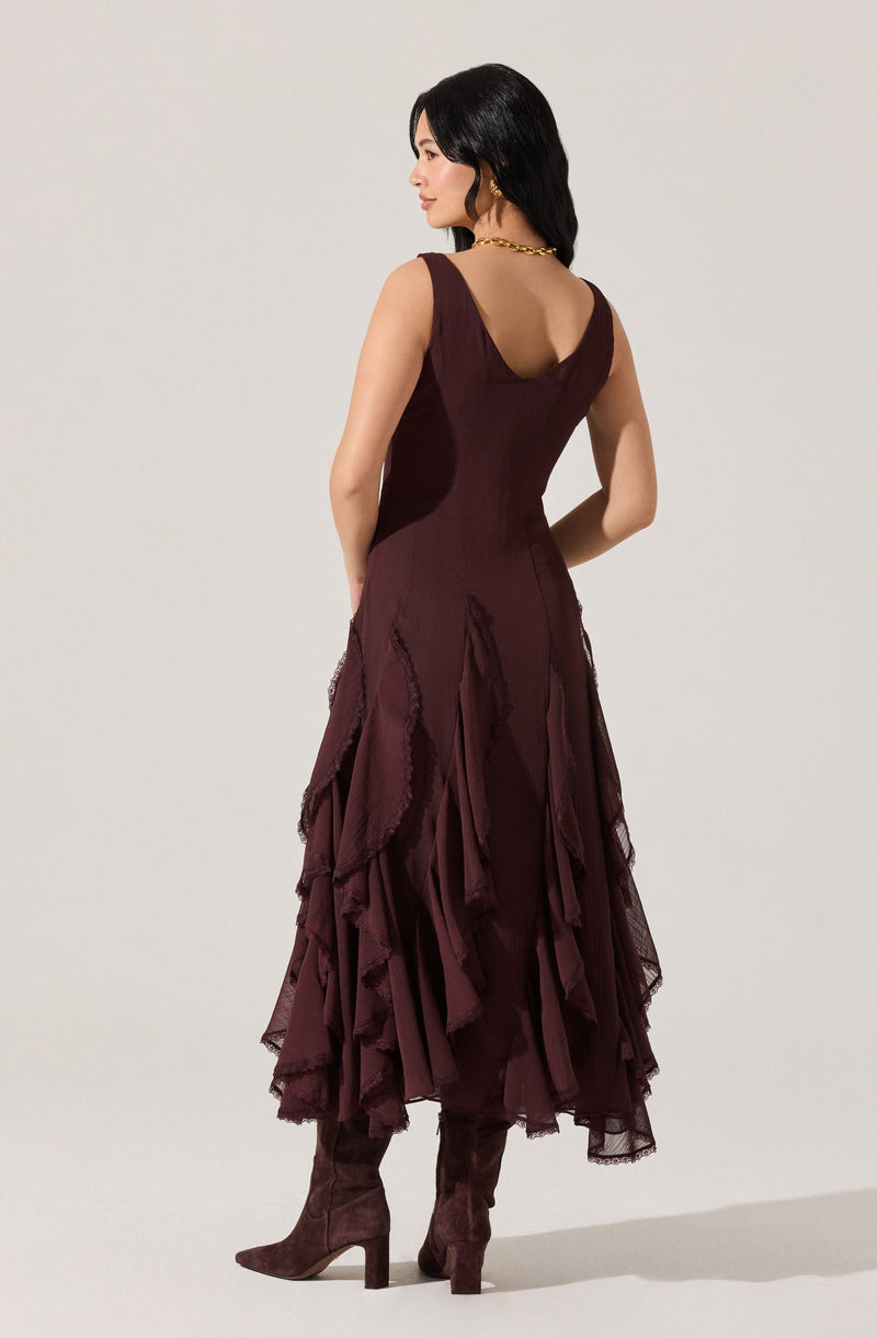 A woman is wearing a sleeveless, deep burgundy dress with a fitted bodice and a V-shaped neckline. The skirt is long and layered with ruffled, uneven panels made of sheer, textured fabric, creating a voluminous and dramatic look. She is also wearing matching high-heeled, knee-high boots in the same burgundy color.