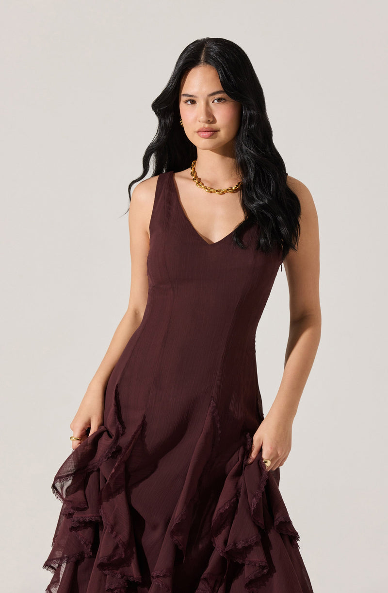 A woman is wearing a sleeveless, deep burgundy dress with a V-neckline. The dress has a fitted bodice and features layered ruffle details at the skirt. She accessorizes with a chunky gold chain necklace and gold hoop earrings.