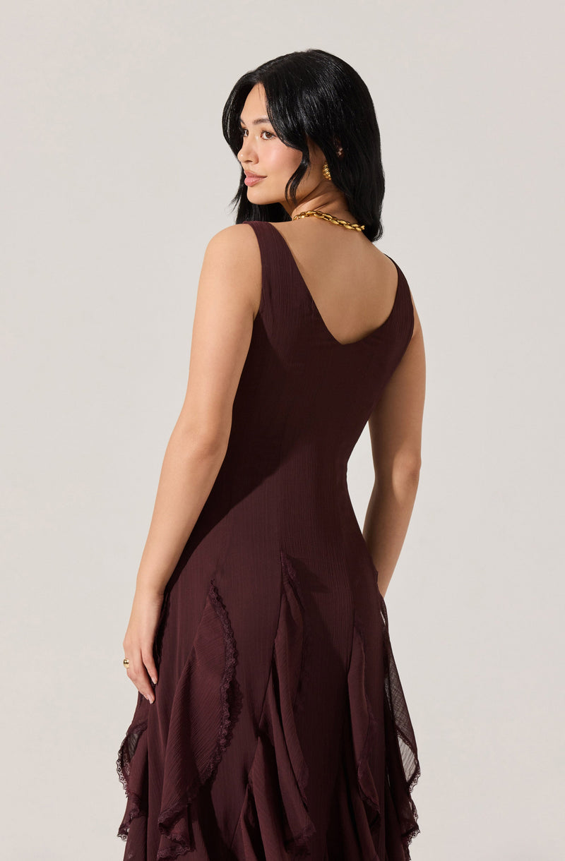A woman is wearing a sleeveless, dark burgundy dress with a fitted bodice that has a scoop neckline in the back. The skirt features layered, ruffled fabric with textured, lace-like details. She accessorizes with a chunky gold chain necklace and small gold earrings.