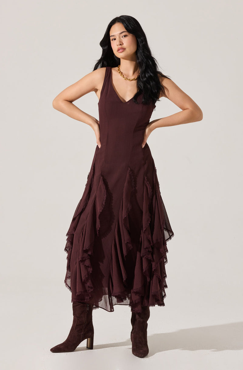 A woman is wearing a sleeveless, deep burgundy dress with a V-neckline. The dress features an asymmetrical, layered ruffled skirt made of a sheer, textured fabric. She has paired the dress with matching burgundy knee-high boots and accessorized with a chunky gold chain necklace.