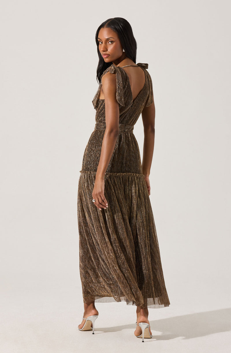 A woman is wearing a long, metallic gold dress with a shimmer effect. The dress features ruffled shoulder details and a fitted waist, with the skirt flowing to the floor in a pleated style. She is also wearing white high-heeled sandals.