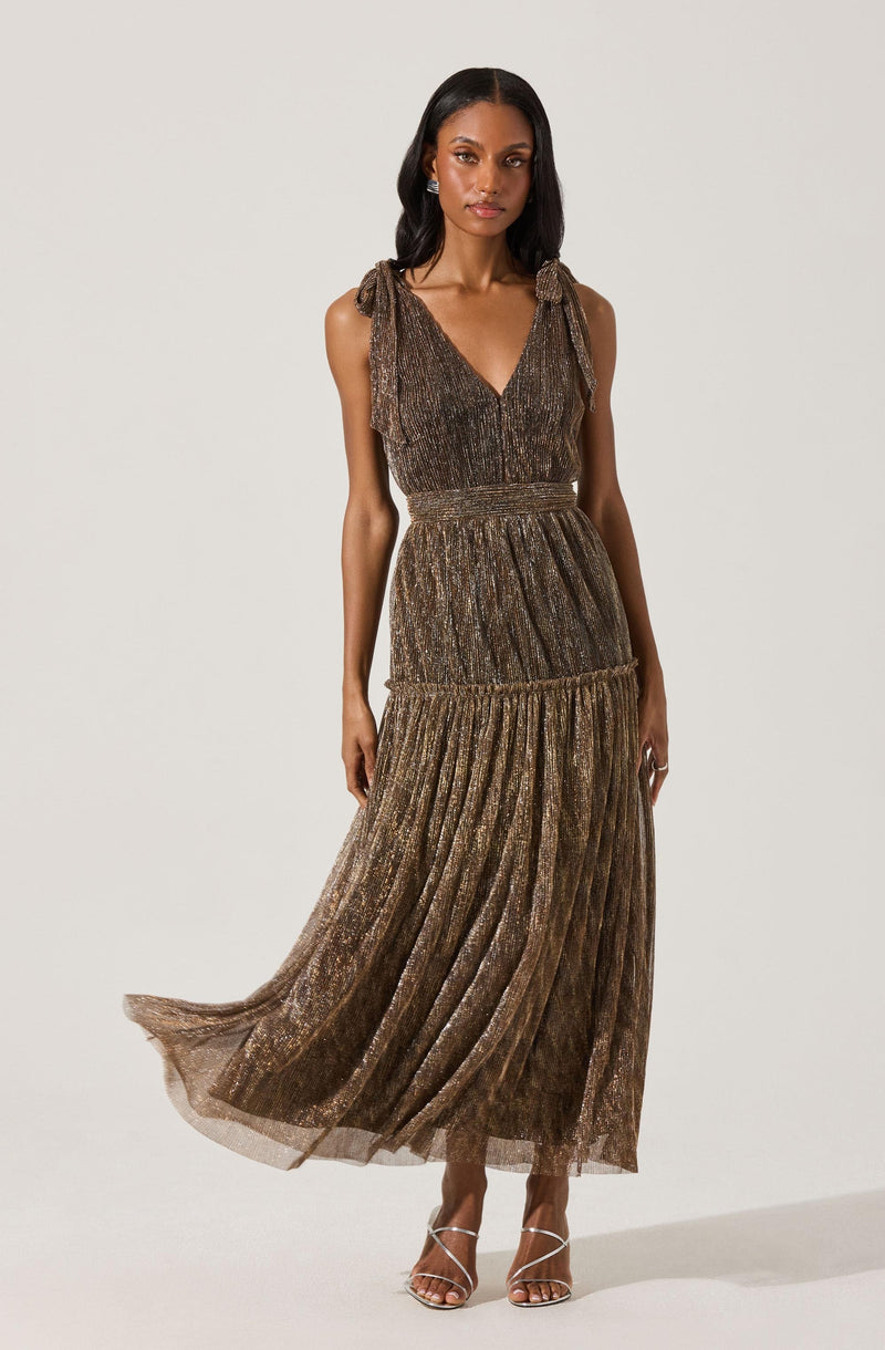 A woman is wearing a sleeveless, full-length dress with a V-neckline and tied shoulder straps. The dress features horizontal bands of fabric with a metallic, shimmery, and textured appearance, creating a layered look. The skirt is flowing and pleated. She is also wearing open-toe, strappy high heels.