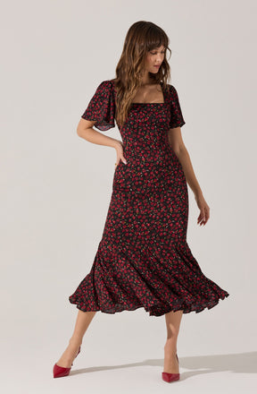 A woman is wearing a long, black dress with a red and pink floral pattern. The dress has short, puffy sleeves made of a sheer fabric, and a fitted bodice with a square neckline. The skirt is flowing and reaches mid-calf, with a ruffled hem and a slight asymmetrical hemline. She is also wearing red high-heeled shoes.