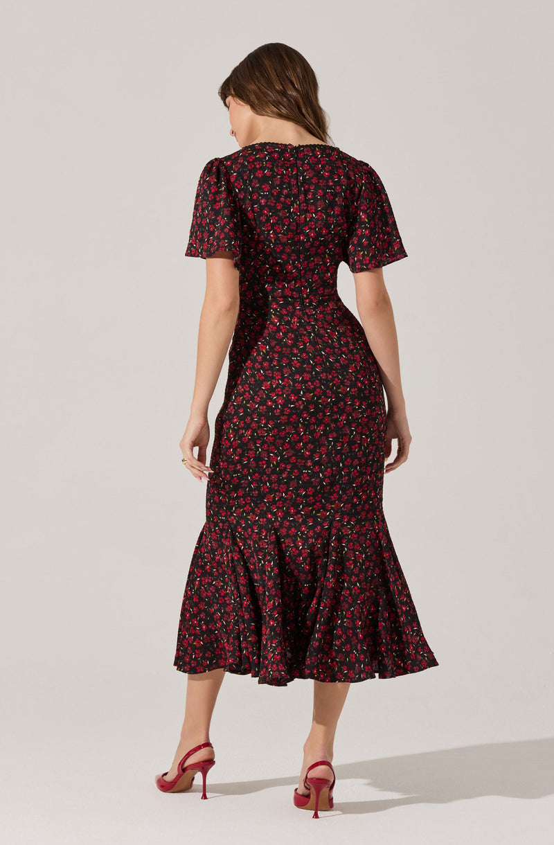 A woman is wearing a black dress with a red floral pattern. The dress has short, puffed sleeves and features a fitted waist with a gathered, ruffled hem at the bottom. She is also wearing red high-heeled shoes.
