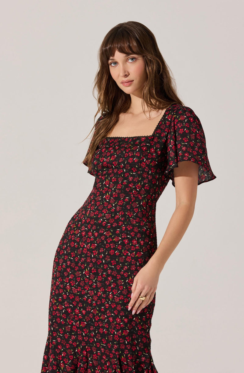 A woman is wearing a black dress with a small red floral pattern. The dress has a square neckline, short, ruffled sleeves, and a fitted waist. The fabric appears lightweight and slightly sheer on the sleeves.
