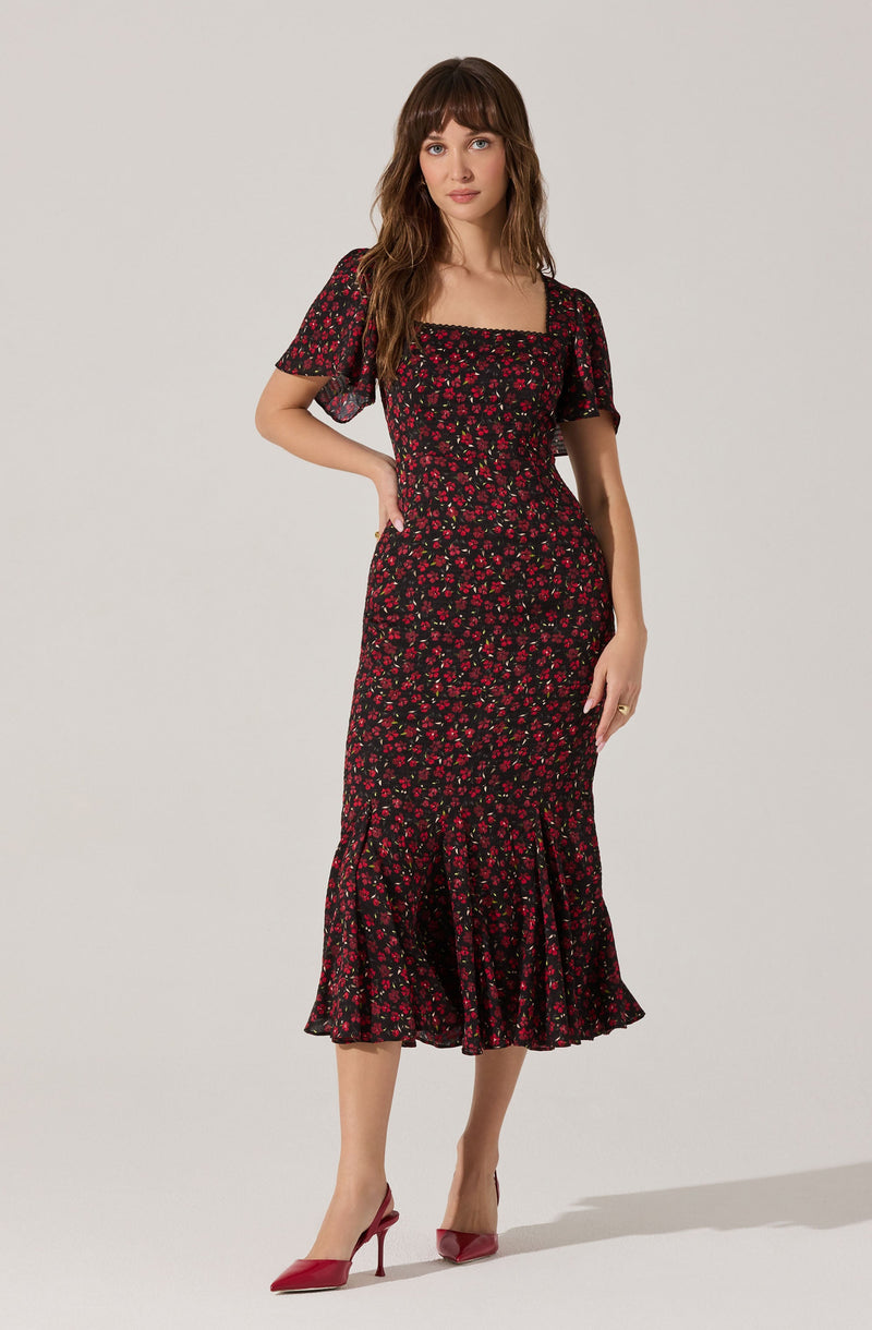 A woman is wearing a black dress with a red floral pattern. The dress has short, semi-sheer puff sleeves and a smocked bodice. The skirt is fitted through the hips and flares out below the knees with a ruffle hem. She is also wearing red high-heeled shoes.