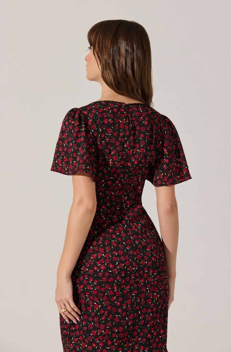 A woman is wearing a black dress with a red floral pattern. The dress has short, slightly puffed sleeves and a fitted waist. The fabric appears lightweight and slightly sheer, especially on the sleeves. The floral design covers the entire dress, creating a vibrant, elegant look.